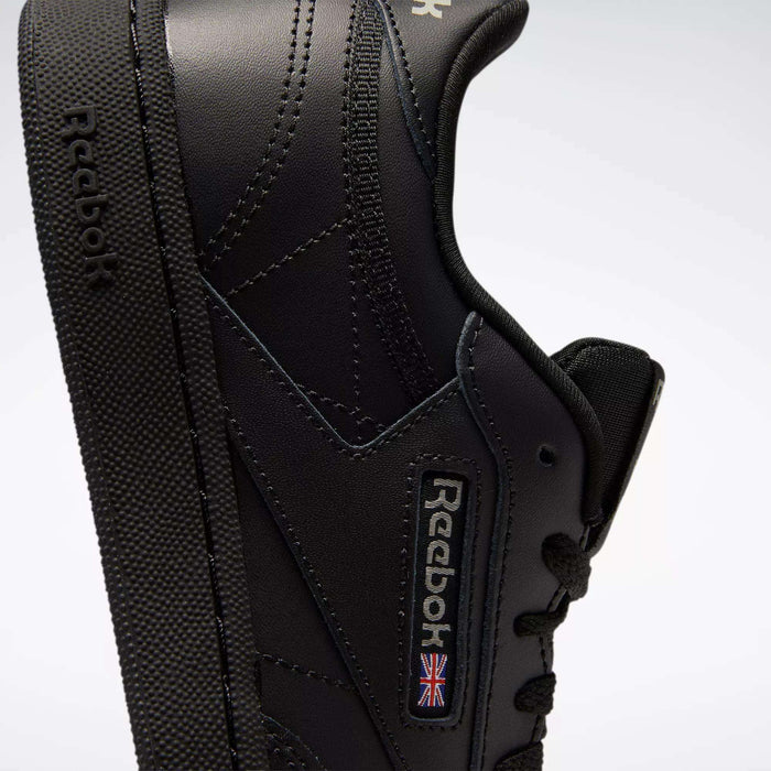 Close-up of Reebok Kids' Club C Shoes - Grade School in black, featuring a side view with visible stitching, soft leather uppers, textured sole, and Reebok branding with a small Union Jack flag.