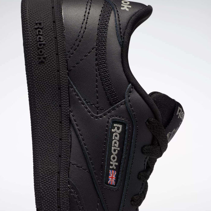 Close-up of the Reebok Kids' Club C Shoes for little kids, featuring a black leather upper with the Reebok logo and a small British flag patch on the side. The shoe is slightly tilted to show its side profile and textured sole.