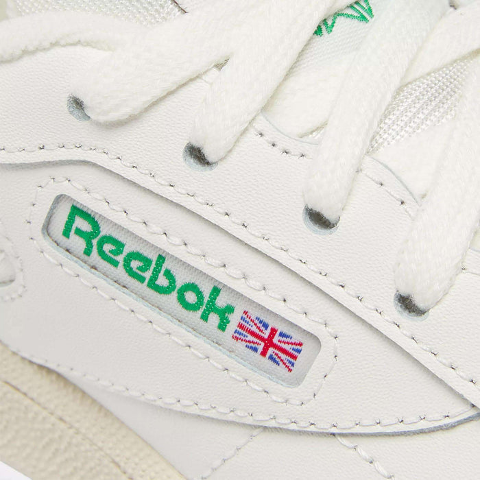 Close-up of the Reebok Kids' Club C Shoes - Preschool in white, featuring a green Reebok logo, Union Jack flag patch, white laces, and classic stitching—an iconic '80s-inspired style from Reebok.