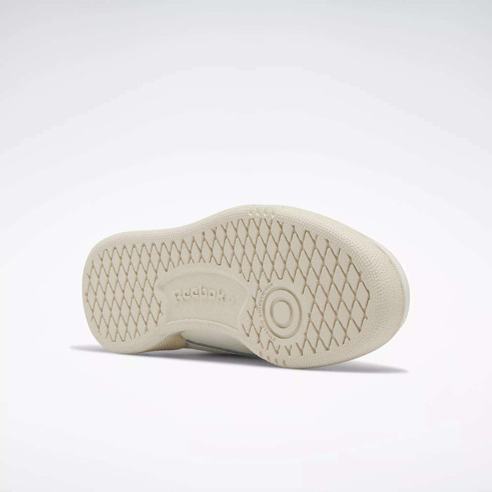 The image shows the sole of a beige Reebok Kids' Club C Shoe - Preschool with a diamond-patterned tread and the Reebok logo at the center. The clean sole and classic style evoke '80s vibes, with the shoe lying on its side against a plain background.