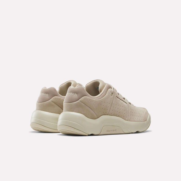 A pair of Reebok DMX Comfort+ Classic walking shoes in beige, featuring chunky off-white soles and crafted from premium leather suede with perforated side details, displayed from the back and side.