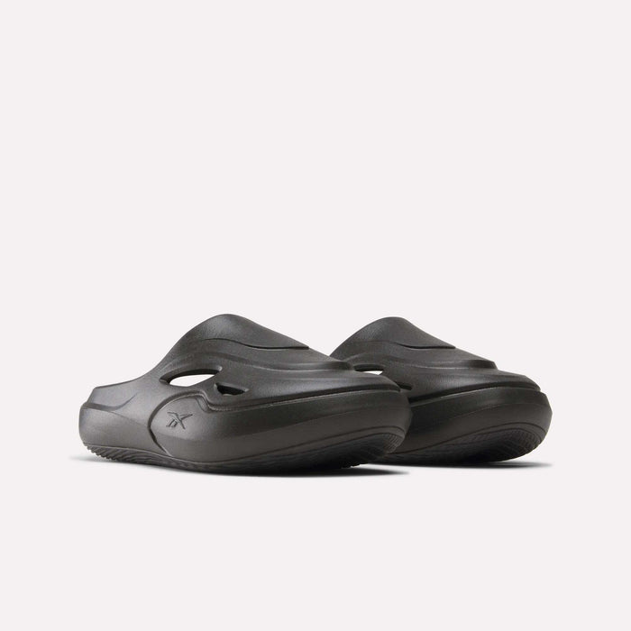 The Reebok DMX Recovery Mules are chunky dark gray molded EVA slip-on slides with side cutouts and grooved soles, shown against a plain light background.