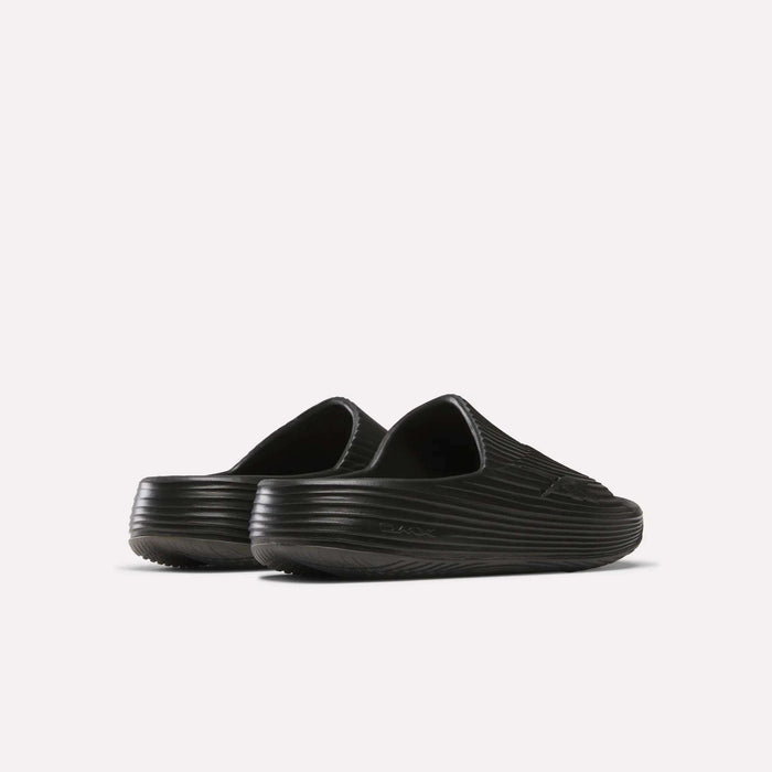 The Reebok DMX Recovery Walking Slides are black slip-on clogs with a ribbed texture, closed toes, and super-soft foam for extra comfort, shown from the back and side on a plain white background.