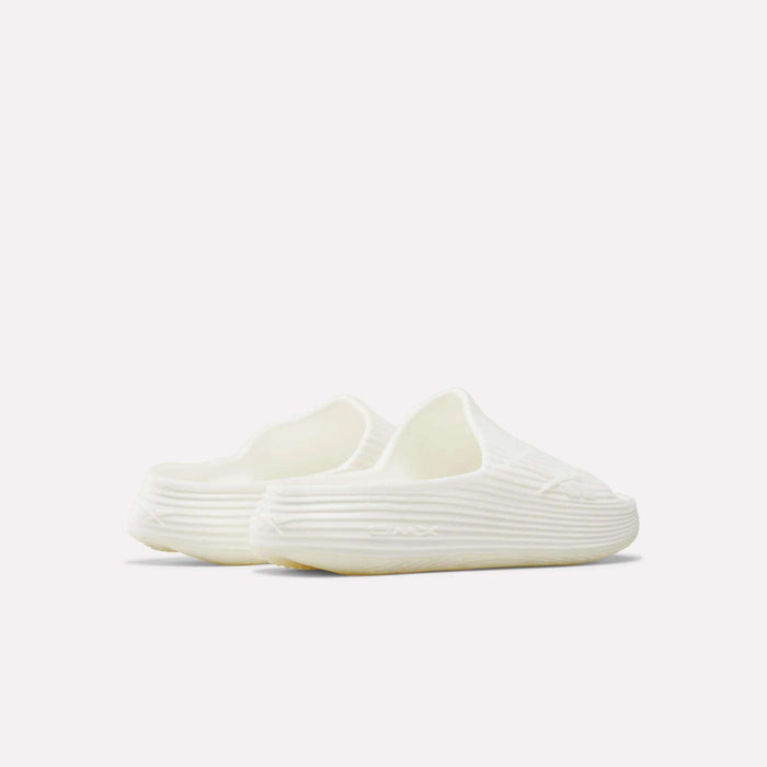 The Reebok DMX Recovery Walking Slides are off-white, slip-on foam clogs with a ribbed texture and rounded toe, inspired by recovery slides, shown from a rear side angle against a plain light background.