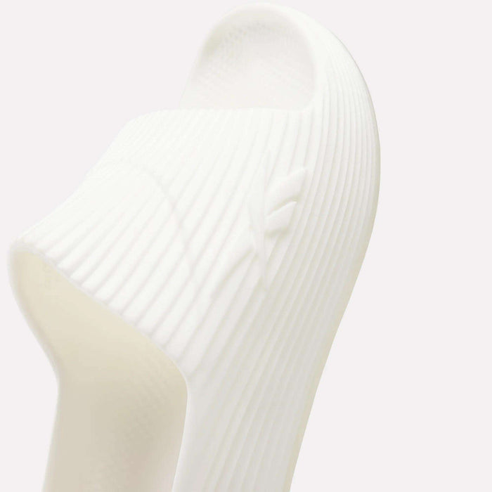 Close-up of white Reebok DMX Recovery Walking Slides featuring ribbed design, subtle embossed logo on the side, and air cushioning, photographed against a light background.