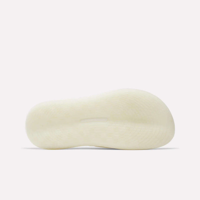 The image shows the bottom view of a Reebok DMX Recovery Walking Slide in white or light beige, highlighting its textured sole with a slightly raised arch. The background is plain and light-colored.