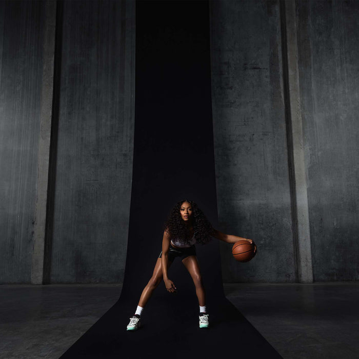 A woman with long curly hair dribbles a basketball on a black runway in front of gray concrete walls, wearing a sports outfit and Reebok Engine A 26 Basketball Shoes designed for energy return and stability, appearing focused and athletic.