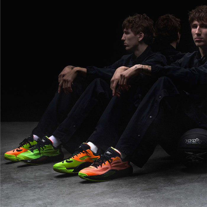 A person in dark clothing sits on the floor, arms on knees, wearing mismatched neon green and orange Reebok Engine A 26 Basketball Shoes. Their reflection and a black Reebok basketball are seen in the background, showcasing style and stability.
