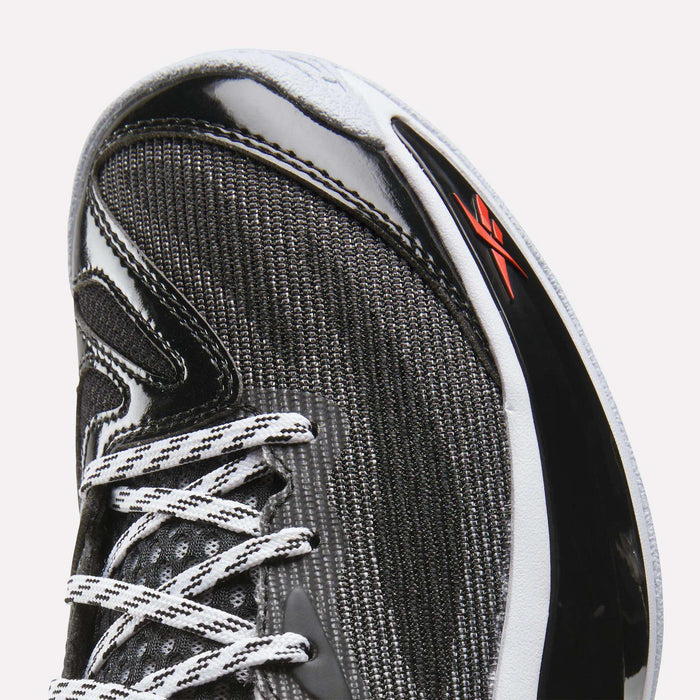 Close-up of the toe and laces of a Reebok Engine A 26 Basketball Shoe in black, white, and gray with a red logo on a glossy black side panel, featuring an engineered mesh upper for enhanced breathability.