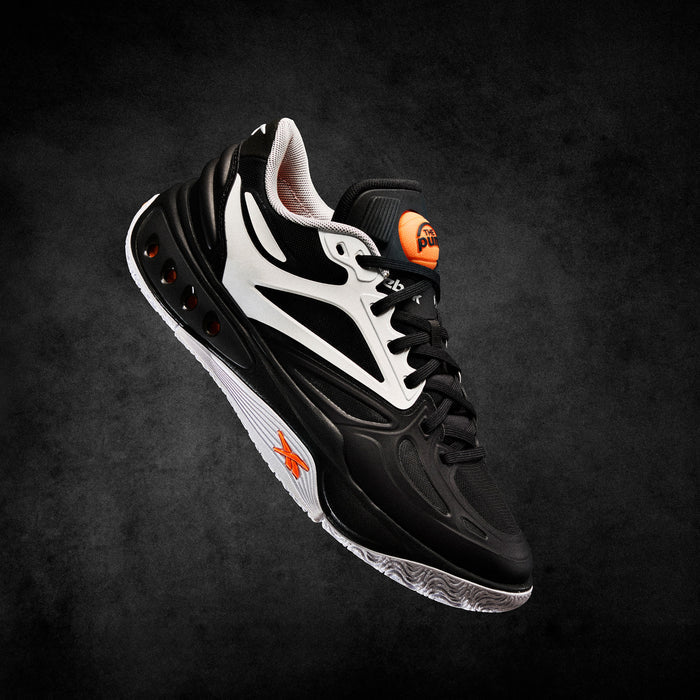 A black and white Reebok Engine A Pump Shoe with striking orange accents floats against a dark textured background, highlighting its side profile, innovative Pump technology, and intricate design details.