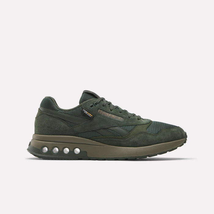 The ERS World Shoes by Reebok are dark green sneakers made with suede and CORDURA mesh. Featuring a chunky sole with circular pods and Energy Return System, these retro running shoes display the Reebok logo on the side against a white background.