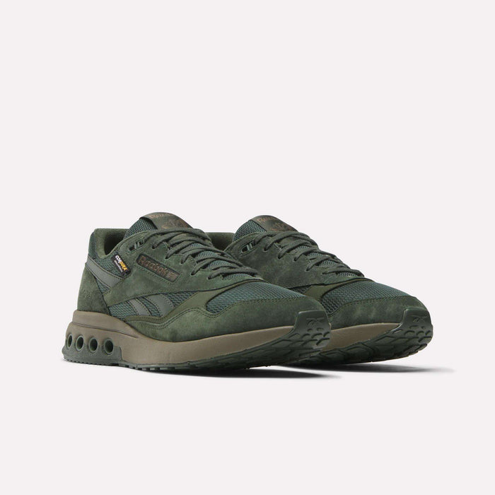 A pair of dark green Reebok ERS World Shoes featuring suede and CORDURA mesh panels, thick soles with circular cutouts, and a rugged tread, shown on a plain white background.