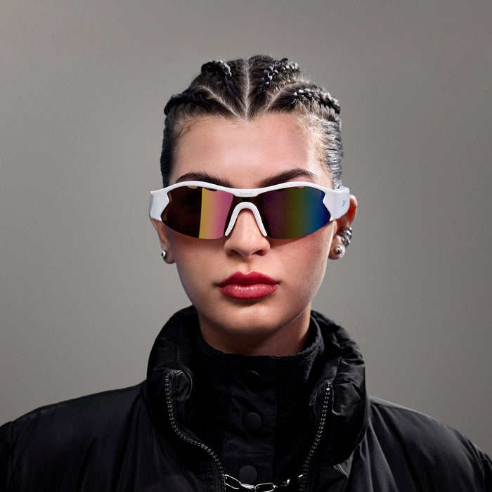 A person with braided hair wearing Reebok Flash Smart Audio Sunglasses with polarized lenses, a black puffer jacket, and bold red lipstick poses against a plain gray background.