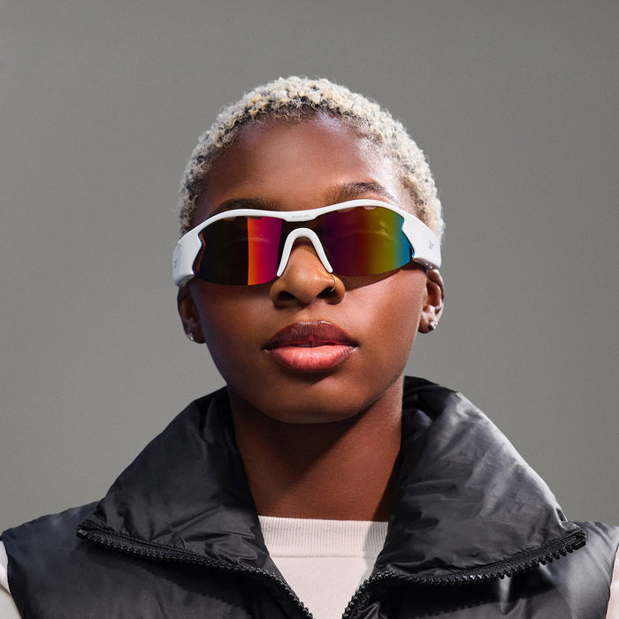 A person with short blond hair wears Reebok Flash Smart Audio Sunglasses featuring polarized lenses and a black puffer vest over a light top, looking straight at the camera against a plain gray background.