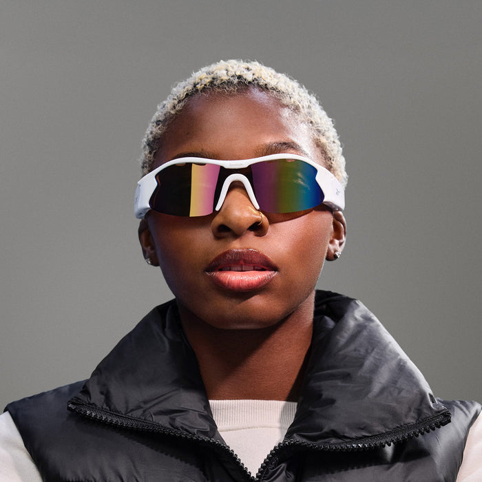 A person with short, bleached hair wears Reebok Flash Smart Audio Sunglasses and a black puffer vest over a light shirt, standing against a plain gray background.