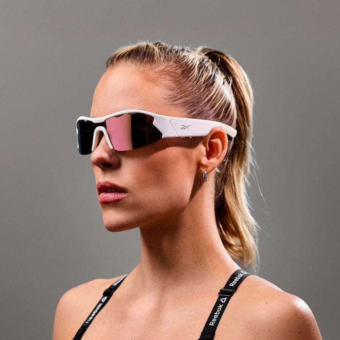 A blonde woman with a ponytail wears Reebok Flash Smart Audio Sunglasses with sporty white polarized reflective lenses and a black strap top, posing against a plain gray background.