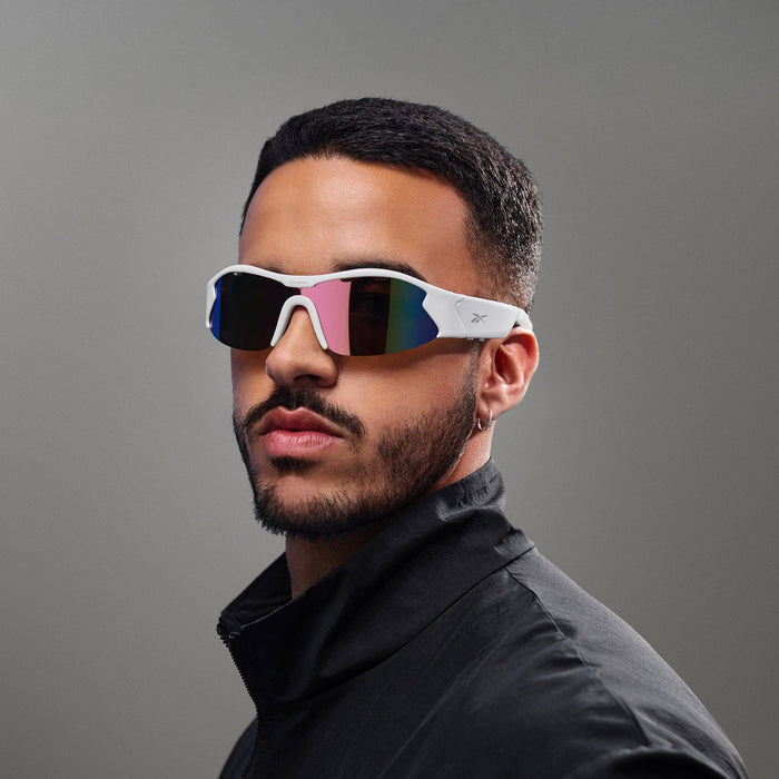 A man with short hair and a trimmed beard wears Reebok Flash Smart Audio Sunglasses with mirrored lenses and a black jacket, posing against a plain grey background.