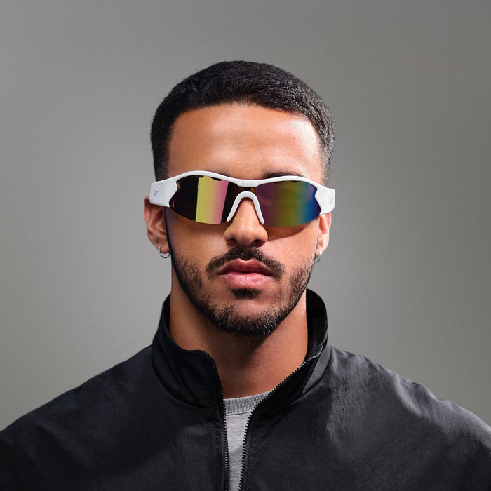 A man with short dark hair and a trimmed beard wears Reebok Flash Smart Audio Sunglasses, a small hoop earring, and a black jacket while posing against a plain gray background.