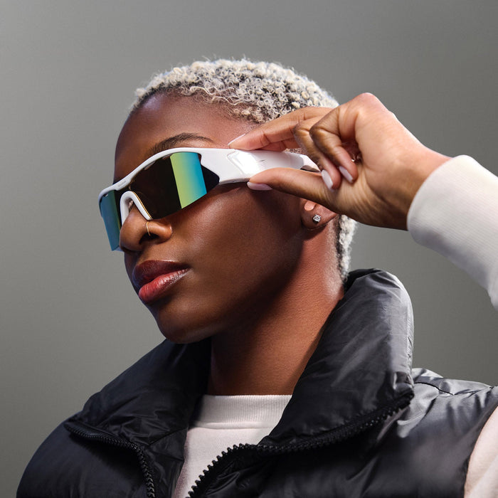 A person with short, bleached blonde hair wears Reebok Flash Smart Audio Sunglasses with polarized lenses and a black puffer vest over a light sweater, touching the side of the sunglasses and gazing confidently against a gray background.
