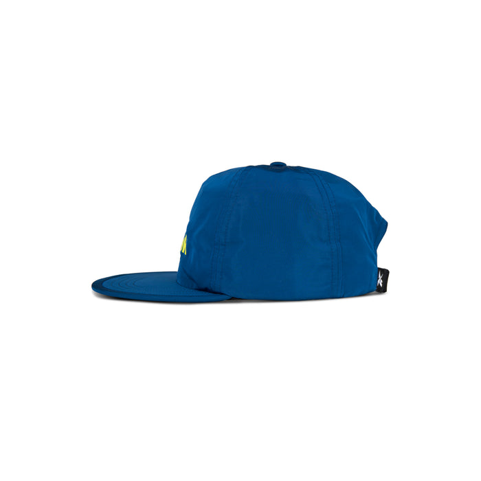The Reebok Flat Brim Running Cap in blue is shown side-on against a white background. This lightweight running hat features moisture-wicking fabric and a small black tag at the back.