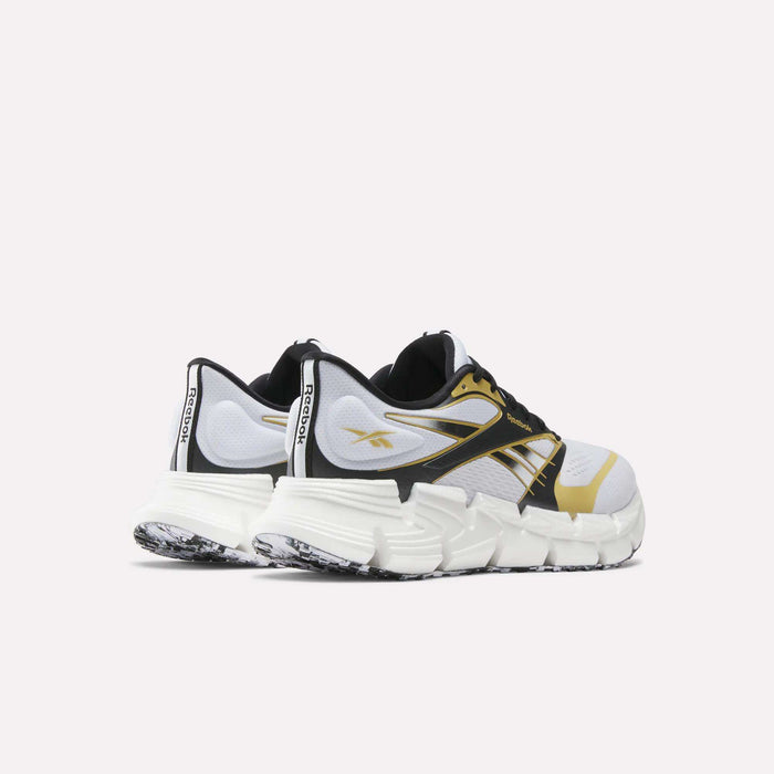 A pair of Reebok FloatZig 2 Running Shoes in white, black, and gold with chunky soles, black laces, a patterned outsole, and energy-returning Superfloat+ cushioning, shown from a rear-side angle on a light background.