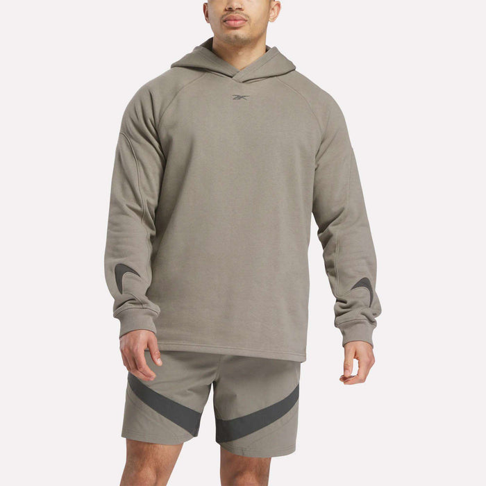A man wears a light brown Reebok French Terry Basketball Hoodie with a small chest logo and matching shorts with dark diagonal stripes, standing against a plain white background.