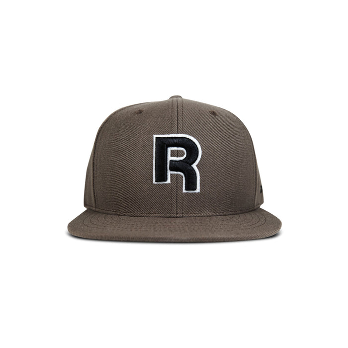 The Reebok Identity Drop R Cap is a brown flat-brim hat with a large black and white "R" embroidered on the front, snapback closure, and is shown against a plain white background.