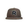 The Reebok Identity Drop R Cap is a brown flat-brim hat with a large black and white "R" embroidered on the front, snapback closure, and is shown against a plain white background.