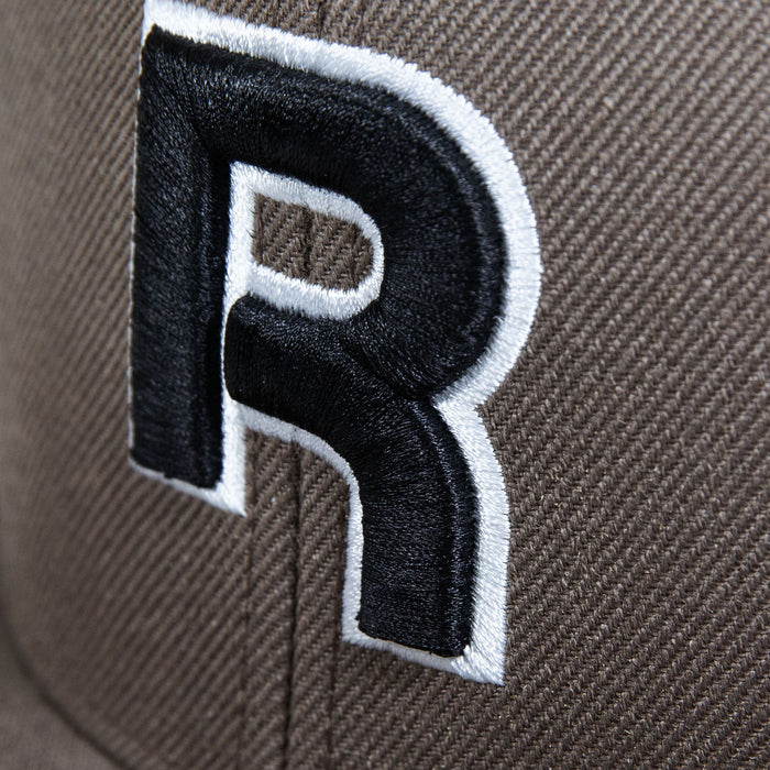 Close-up of Reebok's Identity Drop R Cap in brown, showcasing a large embroidered black "R" outlined in white, with visible stitching, texture, and a flat brim.