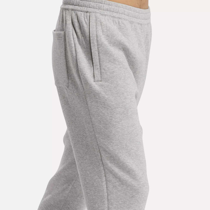 A man models Reebok Men's Active Collective DreamBlend Pants in light gray, featuring an elastic waistband, side pockets, and a relaxed fit, shown from the waist down in profile.