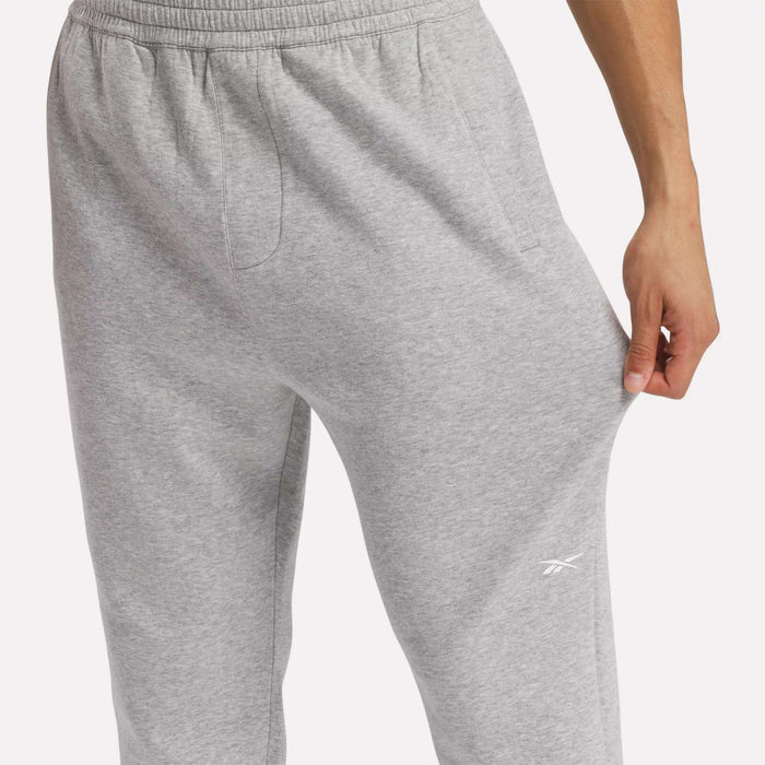 A person wearing light gray Reebok Men's Active Collective DreamBlend Pants tugs at the fabric on their thigh. These tapered pants have an elastic waistband, soft DreamBlend fabric, and a small Reebok logo near the left pocket.