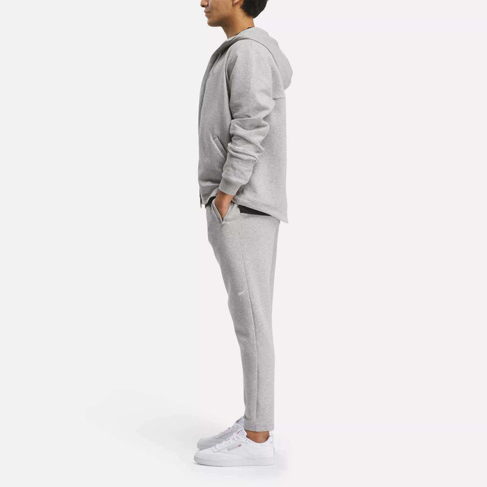 A person stands in profile, hands in pockets, wearing a light gray Reebok Men's Active Collective DreamBlend Pants with a matching hoodie and white sneakers against a plain white background.