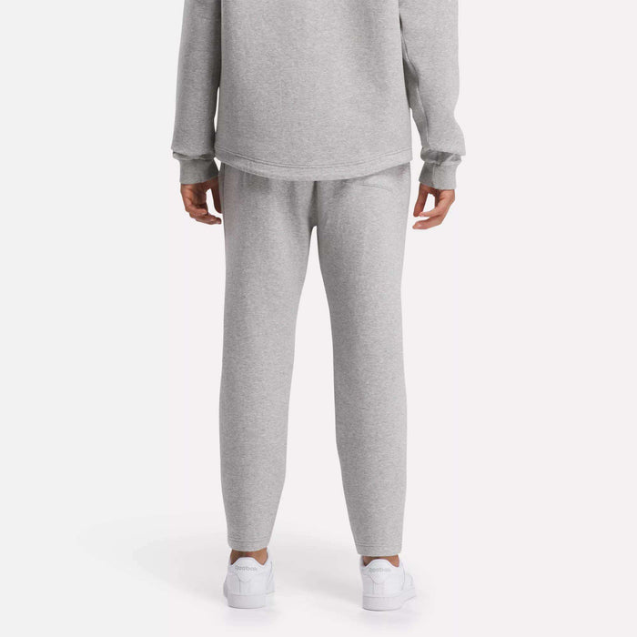 A person is seen from behind in a plain, light setting, wearing a light gray sweatshirt, white sneakers, and Reebok Men's Active Collective DreamBlend Pants with a tapered fit.