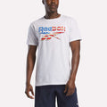 A person wearing a Reebok Men's Americana Graphic T-Shirt in white with a stylized American flag logo, made of soft ringspun cotton, paired with navy shorts, stands against a plain background.