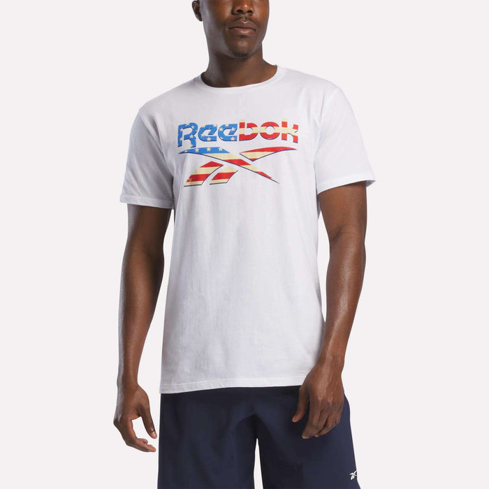 A person wearing a Reebok Men's Americana Graphic T-Shirt in white with a stylized American flag logo, made of soft ringspun cotton, paired with navy shorts, stands against a plain background.