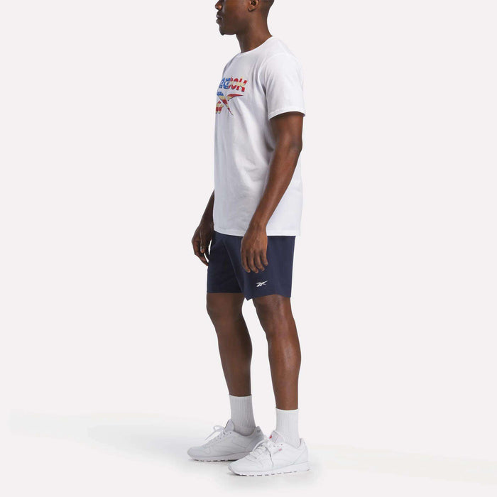 A man poses against a plain white background wearing a Reebok Men's Americana Graphic T-Shirt, navy blue shorts, white socks, and white sneakers.