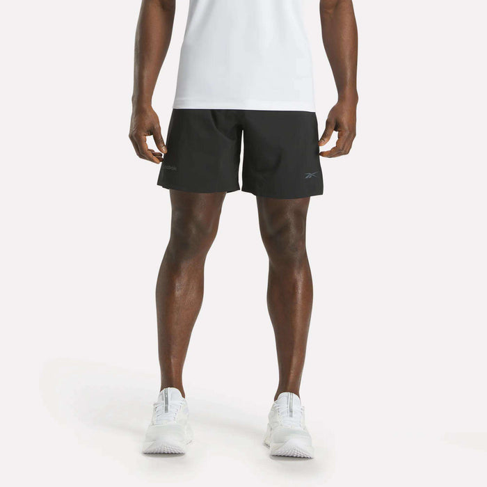 A person is shown from the shoulders down, facing forward against a plain white background, wearing a white shirt, white athletic shoes, and black Reebok Men's Athlete Speed Shorts featuring RBK-DRY technology.