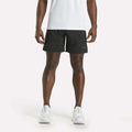 A person is shown from the shoulders down, facing forward against a plain white background, wearing a white shirt, white athletic shoes, and black Reebok Men's Athlete Speed Shorts featuring RBK-DRY technology.