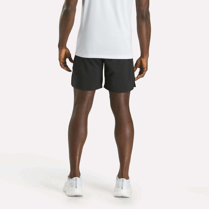 A person stands facing away from the camera, wearing a white T-shirt, Reebok Men's Athlete Speed Shorts in black, and white sneakers against a plain light background.