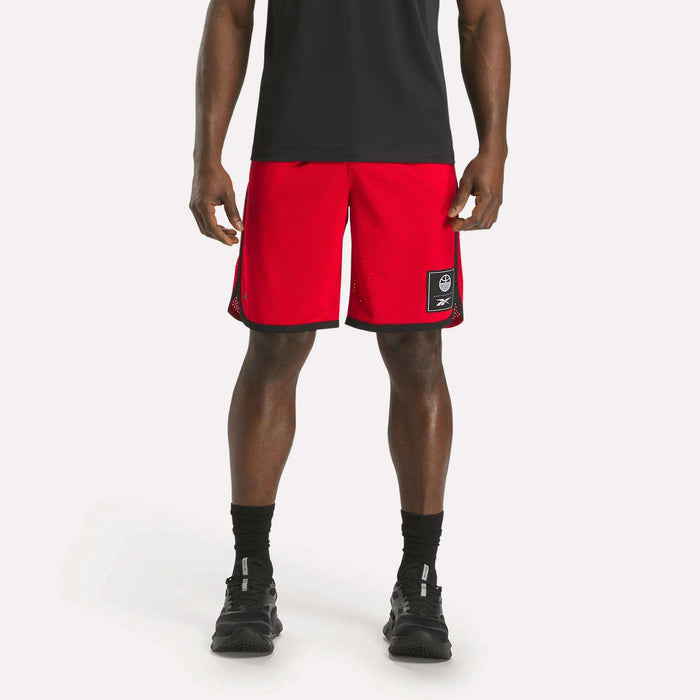 A person wearing a black shirt, Reebok Men's Basketball Mesh Shorts in red with RBK-DRY tech and a patch on the left leg, black socks, and black sneakers stands against a plain white background.