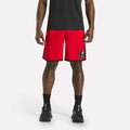 A person wearing a black shirt, Reebok Men's Basketball Mesh Shorts in red with RBK-DRY tech and a patch on the left leg, black socks, and black sneakers stands against a plain white background.
