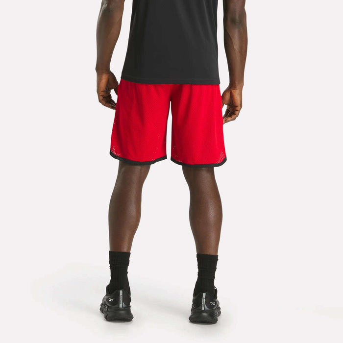 A person stands facing away against a plain white background, wearing a black shirt, black socks, black sneakers, and red Reebok Men's Basketball Mesh Shorts with black trim featuring RBK-DRY technology.