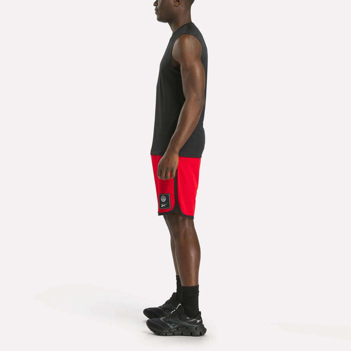 A man stands in profile facing left, wearing a black sleeveless shirt, Reebok Men's Basketball Mesh Shorts, black socks, and black sneakers against a plain white background.