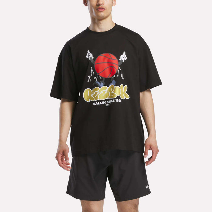 A man in a loose black Reebok Men's Basketball T-Shirt featuring a bold graphic and matching black shorts stands against a plain white background.