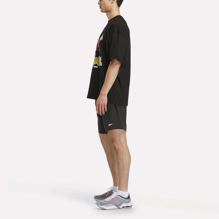 A person in profile wears a black Reebok Men's Basketball T-Shirt, black athletic shorts, and gray athletic shoes, standing against a plain white background.