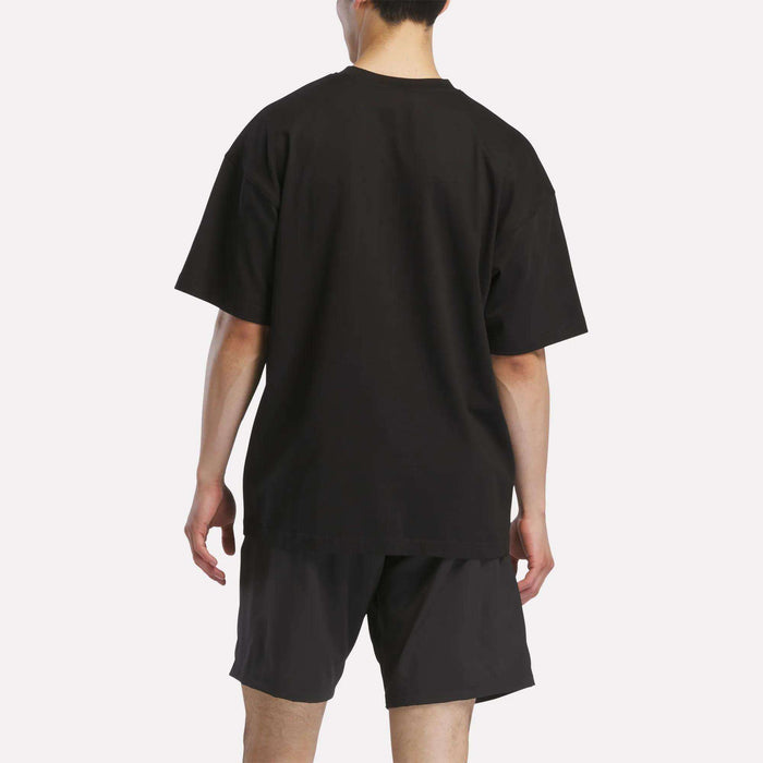 A person faces away from the camera, wearing a loose-fitting black Reebok Men's Basketball T-Shirt and black shorts against a plain white background.