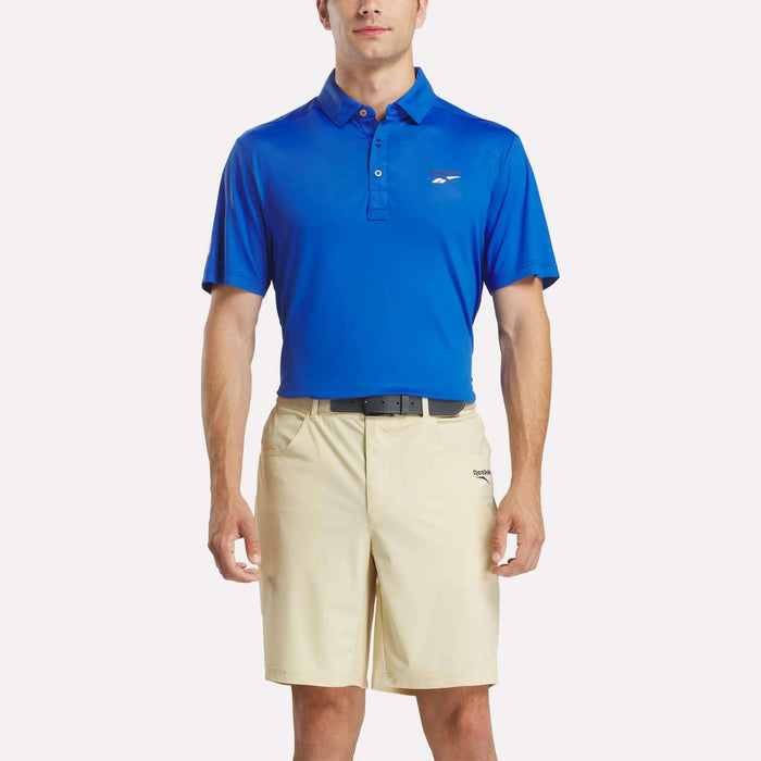 A man stands facing forward against a white background, wearing a Reebok Men's BDC Colorblocked Polo Shirt with RBK-DRY tech, paired with beige shorts and a black belt.