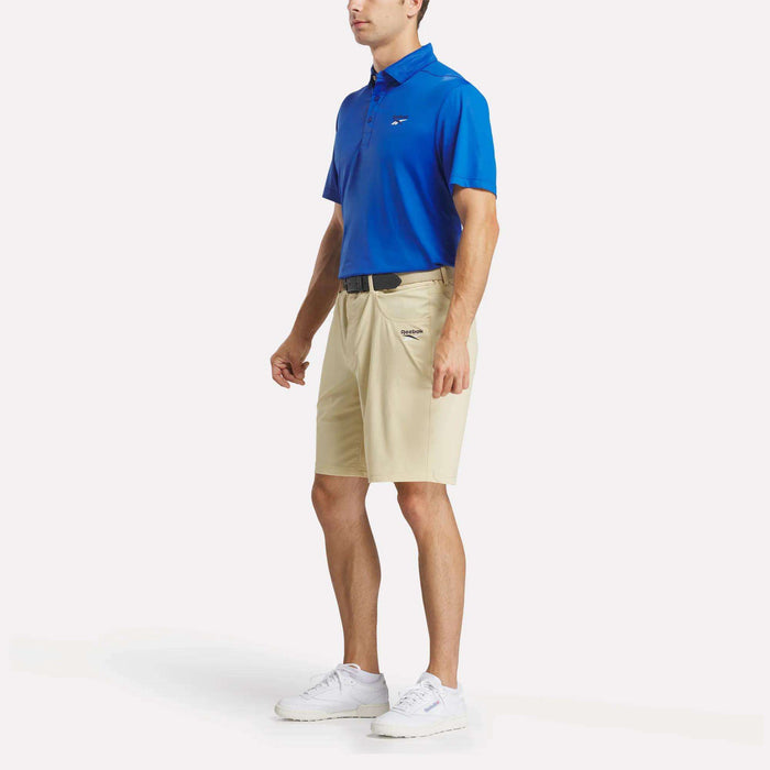 A man wears a blue Reebok Men's BDC Colorblocked Polo Shirt, beige shorts with a black belt, and white sneakers. He stands slightly turned to the side against a plain white background.