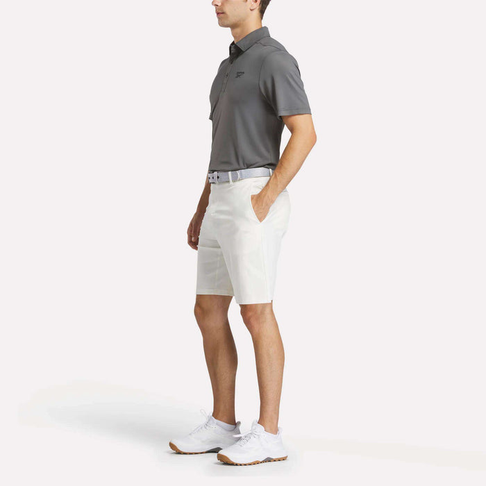 A man stands with hands in his pockets, wearing a gray Reebok Men's BDC Colorblocked Polo Shirt, white shorts, a belt, and white athletic shoes against a plain white background.