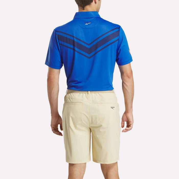 A man stands with his back to the camera wearing a blue Reebok Men's BDC Colorblocked Polo Shirt and light beige shorts.